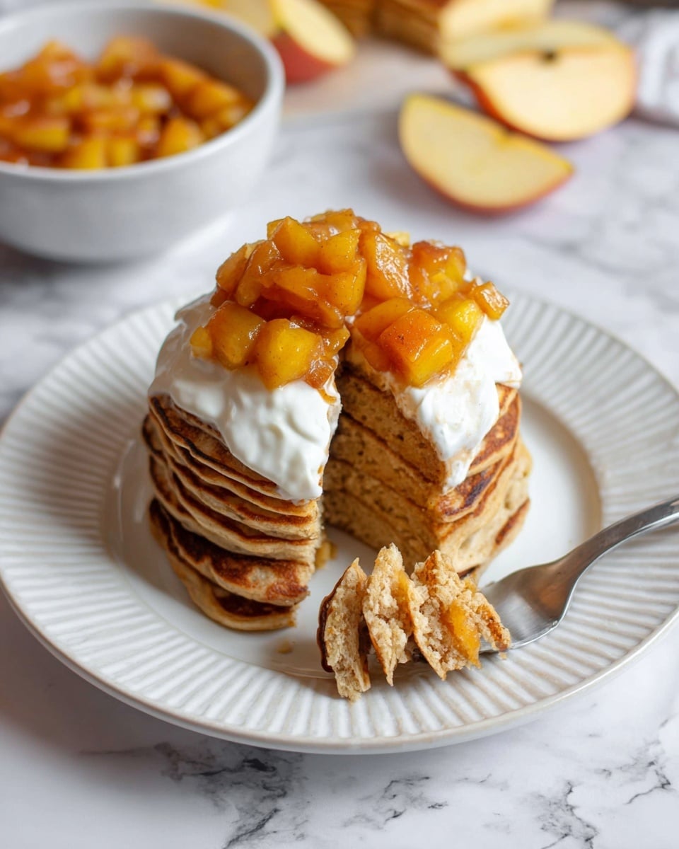 A stack of seven golden brown pancakes sits in the center of a white plate with a rippled edge, topped with a thick layer of white cream and a heap of glossy, orange-brown cooked apple cubes gently spilling onto the cream. A small wedge has been cut from the stack, with a silver fork lifting a bite that shows the soft, fluffy pancake layers inside. In the background, a white bowl filled with more cooked apple cubes and a few sliced pale yellow apples lie on a white marbled surface, enhancing the warm colors of the dish. Photo taken with an iphone --ar 4:5 --v 7