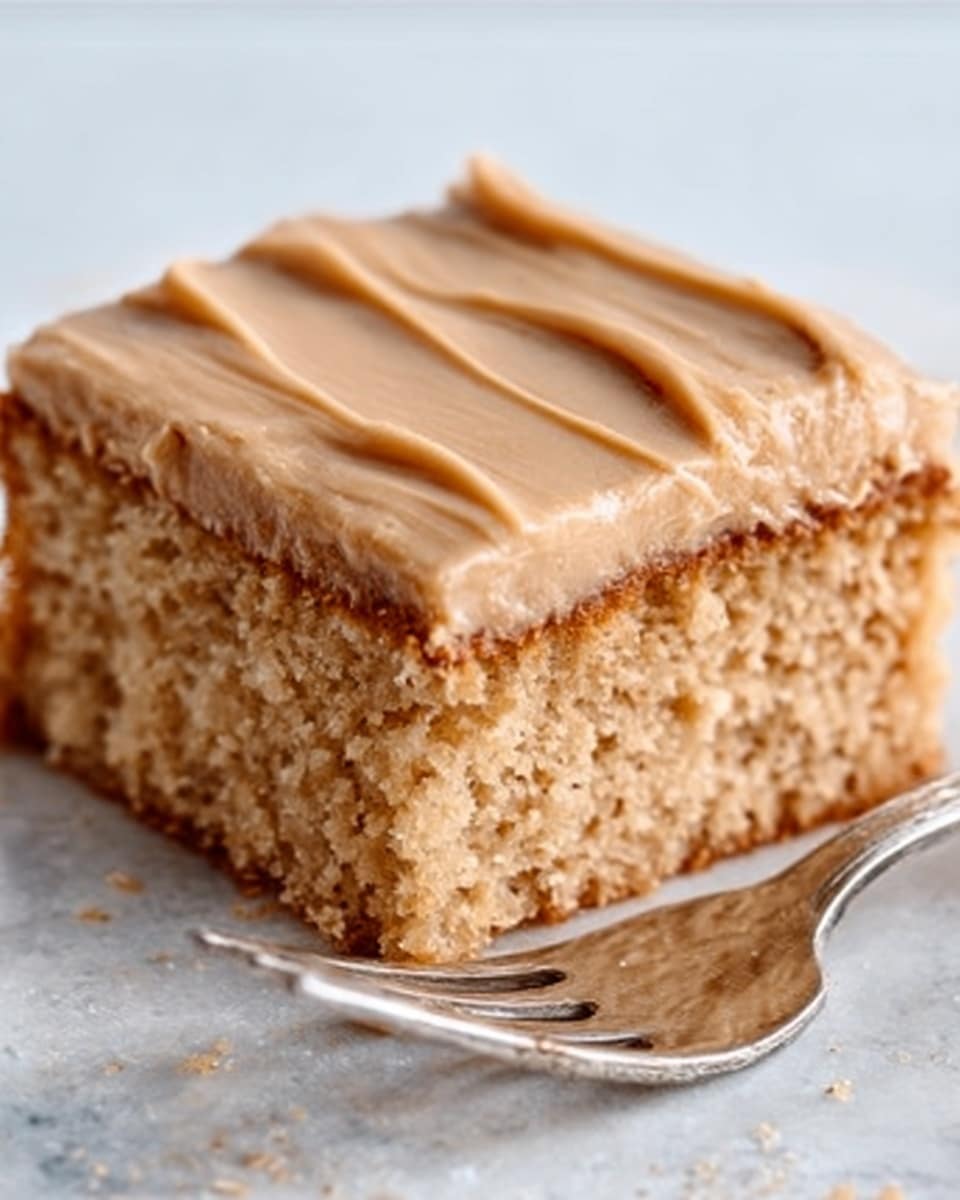 The image shows a square piece of cake with two layers. The bottom layer is light brown and looks soft and crumbly, while the top layer is thick and smooth with light brown frosting spread evenly with four soft ridges on the surface. The cake is placed on a silver fork that lies on a white marbled surface. The photo is clear and close up, showing the cake’s texture well. photo taken with an iphone --ar 4:5 --v 7