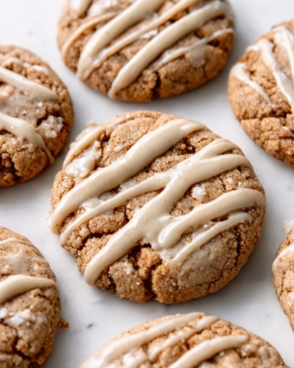 The image shows a close-up of several round, soft cookies with a slightly cracked surface, revealing a chewy texture. Each cookie is topped with light beige drizzle that looks smooth and creamy, applied in uneven thin lines across the top. The cookies sit directly on a white marbled surface, with no plate visible. The colors are mostly warm brown tones from the cookies and pale beige from the drizzle. The lighting is bright and natural, highlighting the texture of the cookies and the shine of the drizzle. Photo taken with an iphone --ar 4:5 --v 7