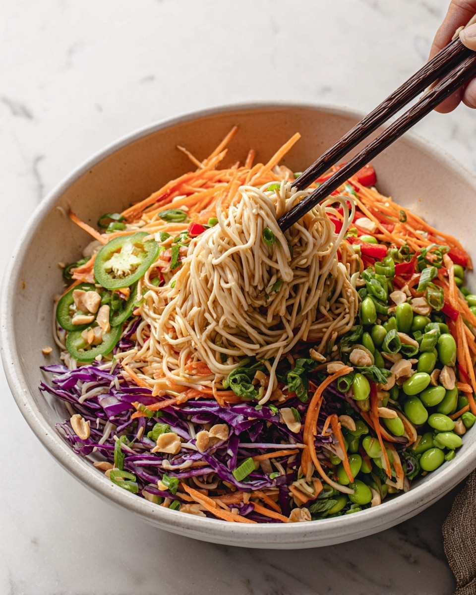 A large white bowl filled with a colorful noodle salad showing about three layers: the bottom layer of thin, light tan noodles, the middle layer of shredded orange carrots and thin strips of red bell pepper, and the top layer scattered with bright green edamame beans, chopped green onions, chopped peanuts, and fresh cilantro leaves. Purple cabbage slices are mixed throughout the salad, adding pops of violet. On the side inside the bowl is a half lime with small cilantro pieces on it, resting on a bed of fresh cilantro leaves. The bowl is set on a white marbled surface with some small bowls containing black and white sesame seeds and additional chopped peanuts and cilantro nearby. The salad looks fresh, crunchy, and vibrant, photo taken with an iphone --ar 4:5 --v 7