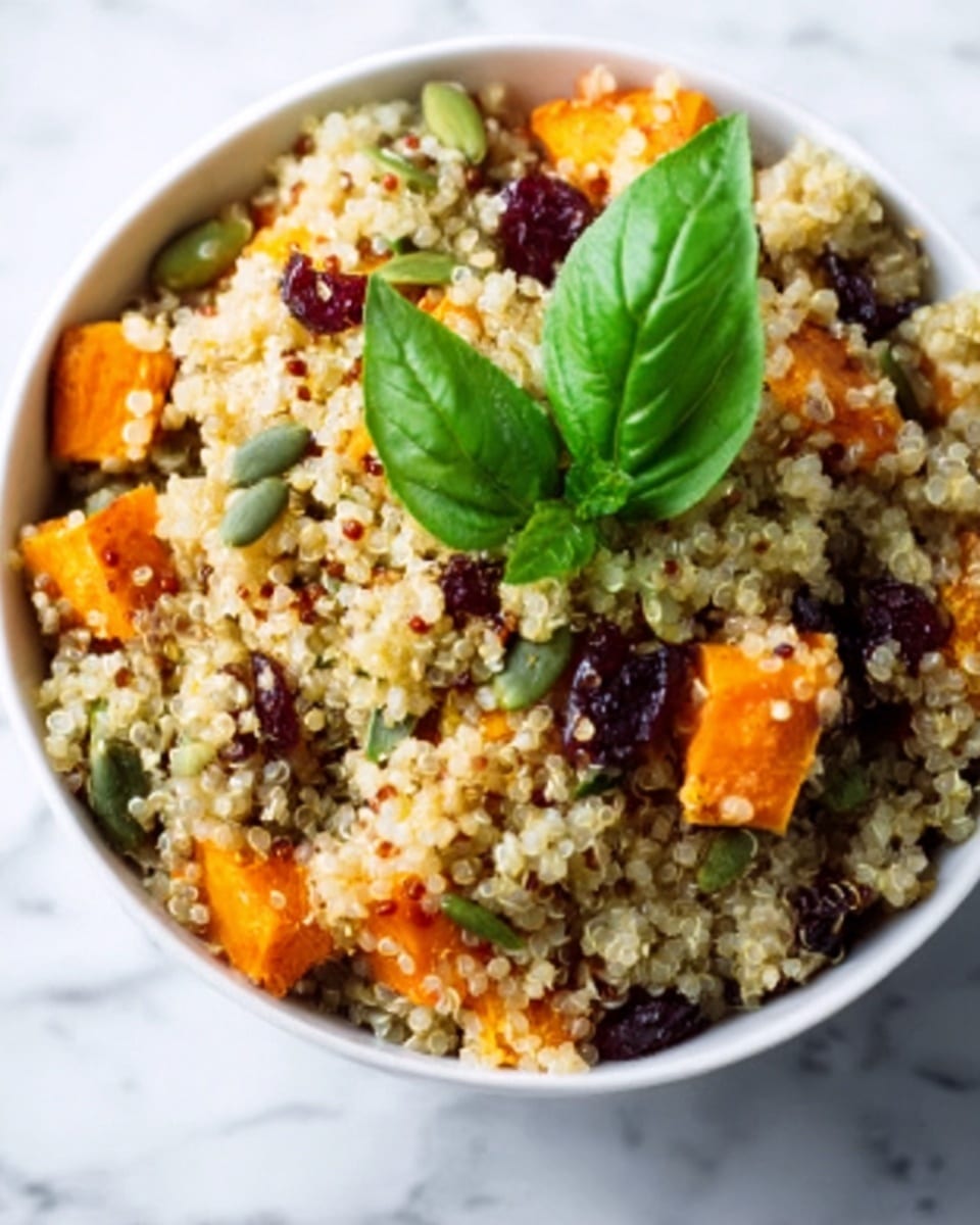 The image shows a close-up of a bowl of quinoa salad placed on a white marbled surface. The bowl is white and filled with multiple layers of ingredients: the base layer consists of fluffy, light beige quinoa grains. Scattered throughout are bright orange sweet potato chunks, deep red dried cranberries, and green pumpkin seeds. On top, there is a fresh, vibrant green basil leaf positioned in the center, adding a pop of color. The textures in the bowl range from soft quinoa to firm sweet potato and crunchy seeds, creating a colorful and fresh look. Photo taken with an iphone --ar 4:5 --v 7