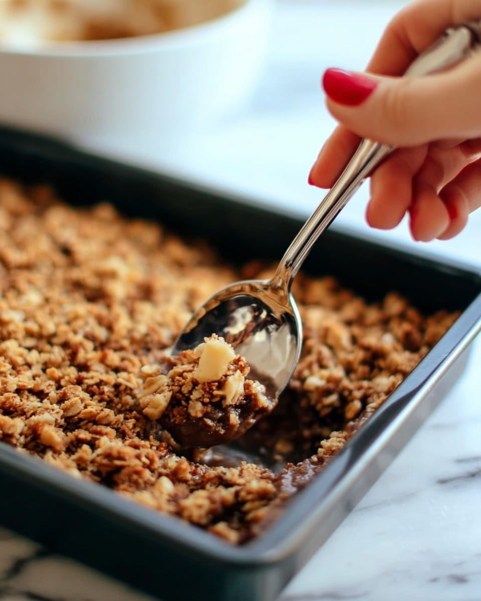 A close-up image showing a woman's hand with red nail polish holding a shiny silver spoon, scooping into a rectangular black baking pan filled with a crumbly, golden brown oat and nut topping, which has a rough and textured surface. The background has a blurred white bowl and a white marbled texture underneath. photo taken with an iphone --ar 4:5 --v 7