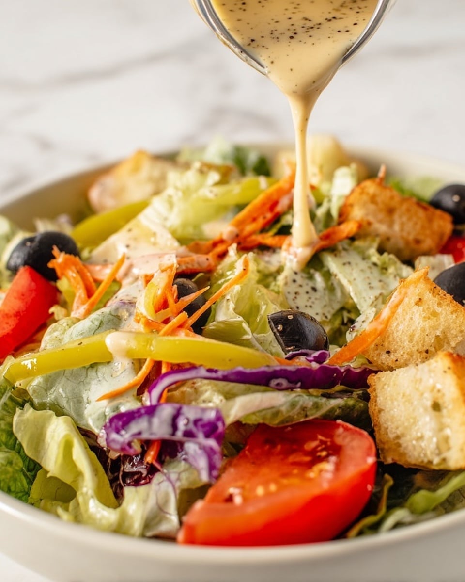 The image shows a fresh salad with multiple layers: the bottom has light green leafy lettuce mixed with some purple cabbage and thin orange carrot strips, scattered black olives sit on top, and slices of red tomato rest around the edges. There are some yellowish-green pepperoncini peppers among the salad, and golden brown croutons add crunch on top of the leaves. Creamy, beige dressing with visible black pepper specks is being poured over the salad from above, creating a flowing texture that drips onto the vegetables. The whole dish is set on a white bowl placed on a white marbled surface. photo taken with an iphone --ar 4:5 --v 7