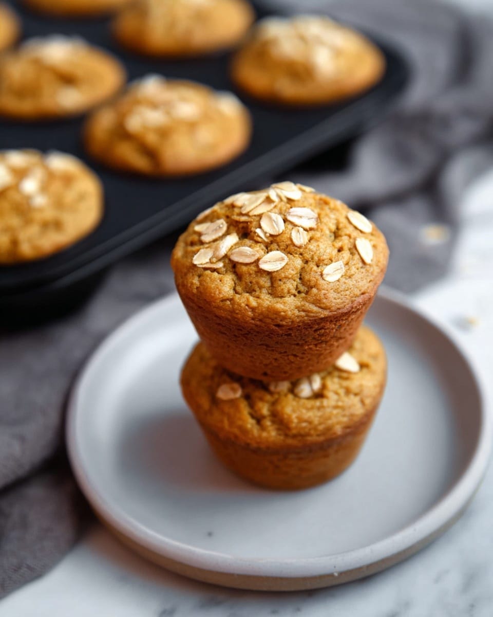 A close-up view of a dark metal muffin tray filled with baked oatmeal muffins. Each muffin rises above its round cup, with a light golden-brown top sprinkled with scattered oat flakes giving a slightly rough texture. The muffins show a soft, uneven surface with small cracks and gentle browning on the edges, creating a warm and fresh look. The background is a white marbled texture. photo taken with an iphone --ar 4:5 --v 7