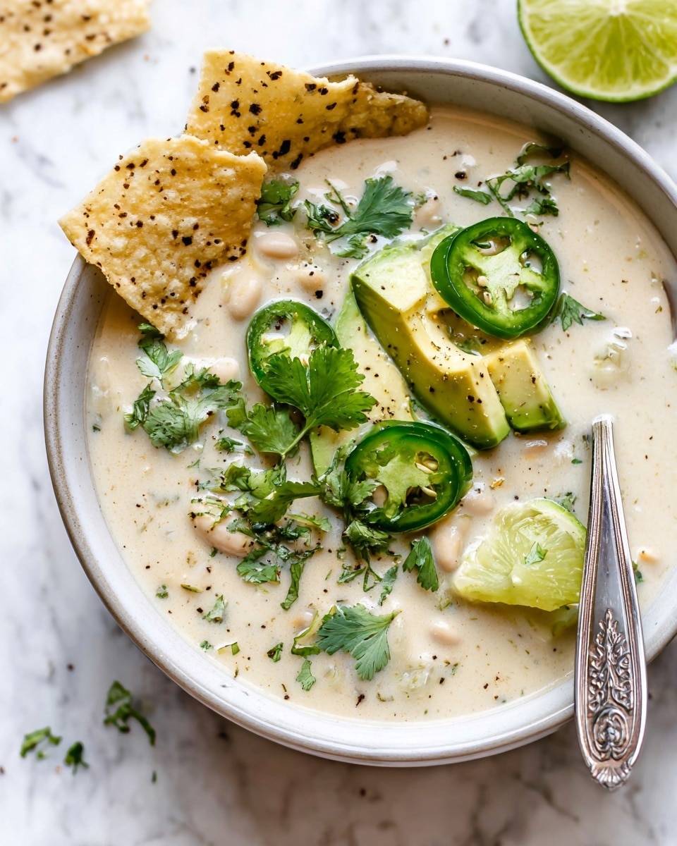 A white bowl filled with creamy white soup showing small chunks of vegetables and beans throughout. On top, there are three light green slices of avocado overlapping each other, along with two round slices of dark green jalapeño peppers. Bright green cilantro leaves are scattered over the surface, with a wedge of light green lime placed on the right side of the bowl. Two pieces of light beige tortilla chips with black seeds are partially dipped in the soup on the left side. A silver spoon with an ornate handle rests inside the bowl on the right. The bowl is placed on a white marbled surface with some scattered black pepper. Photo taken with an iphone --ar 4:5 --v 7