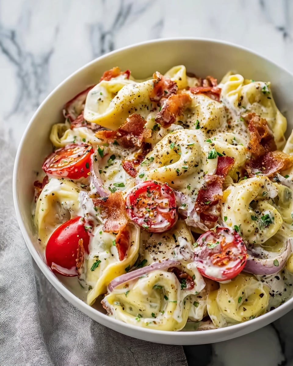 A white bowl filled with creamy tortellini pasta mixed with slices of red cherry tomatoes, crispy pieces of bacon, thin rings of purple onion, and yellow pepper slices, all coated in a thick white sauce. The dish is sprinkled with small green herbs and black pepper flakes, adding texture and color contrast. The bowl is placed on a white marbled surface with a light gray cloth underneath. photo taken with an iphone --ar 4:5 --v 7