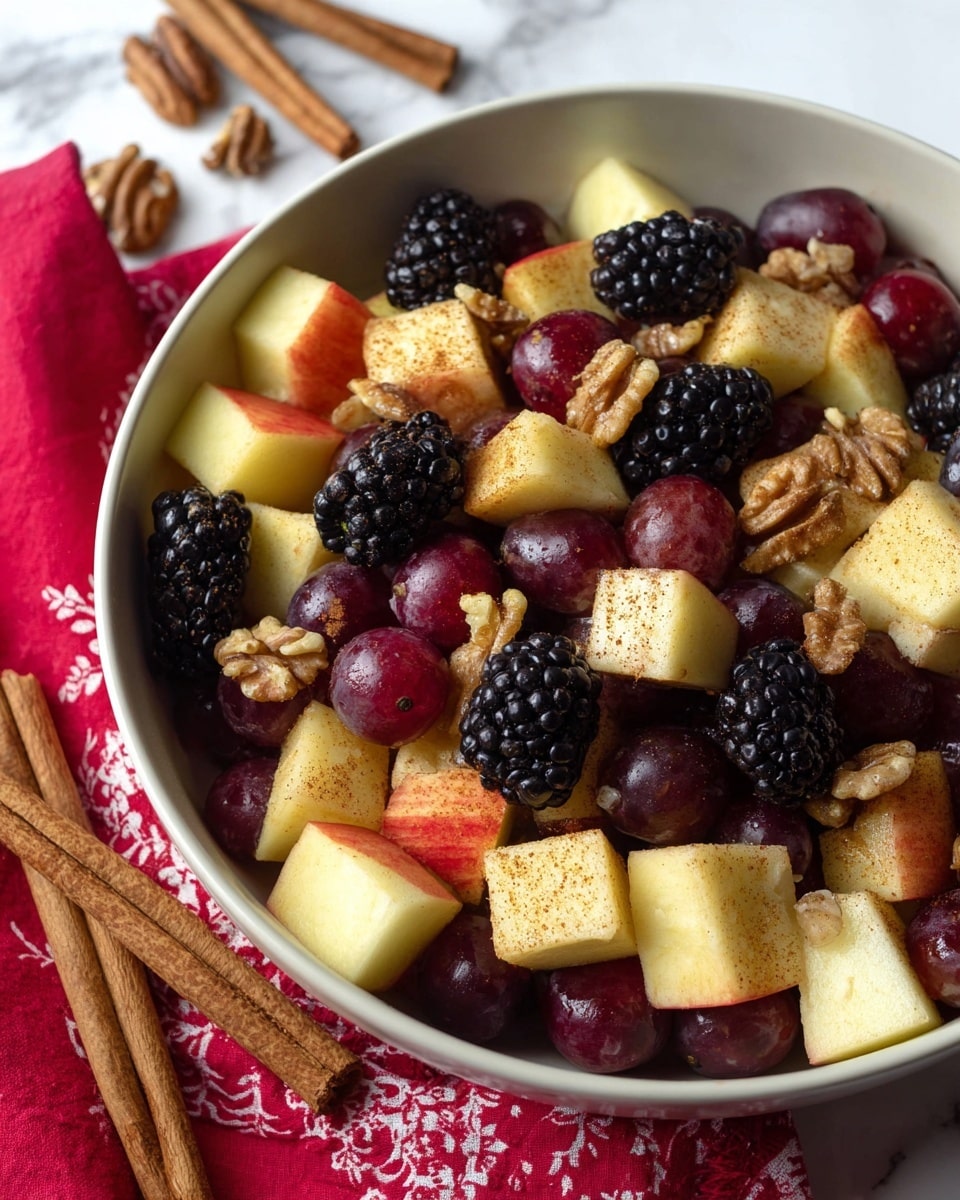 A close-up view of a mixed fruit salad in a white bowl placed on a white marbled surface, showing three main layers: large chunks of pale yellow and red apples with a light dusting of cinnamon on top, shiny deep red grapes scattered throughout, and plump blackberries that add a deep purple-black contrast, all mixed with a layer of rough-textured walnut pieces spread evenly. The background includes a red cloth with cream patterns and two cinnamon sticks lying next to the bowl. Photo taken with an iphone --ar 4:5 --v 7