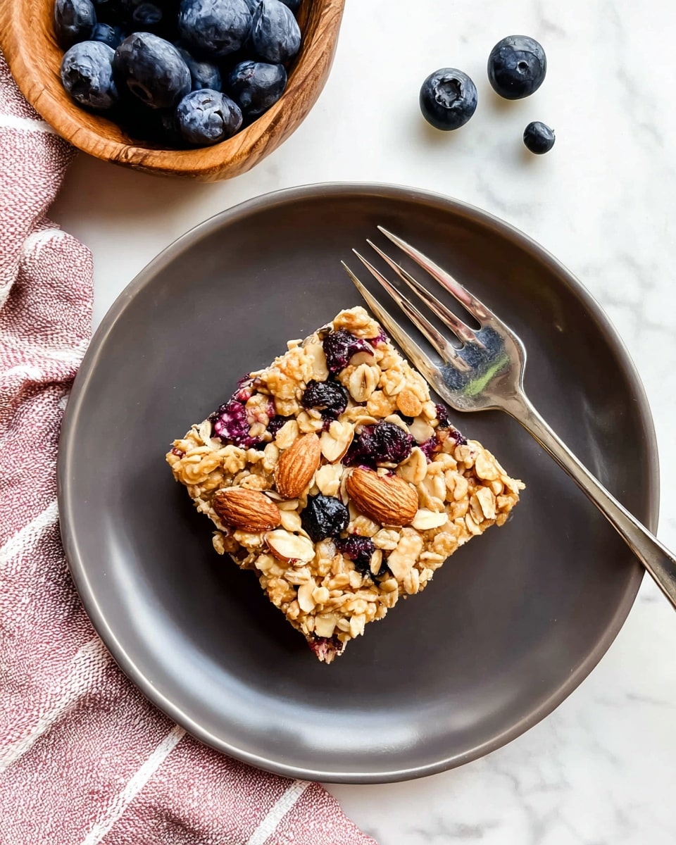 A single square granola bar sits in the middle of a round white plate, showing a texture made of oats, whole almonds, and scattered blueberries that add pops of deep purple and blue color. A silver fork rests diagonally across the plate beside the bar. Around the plate on the white marbled surface, a wooden bowl filled with fresh blueberries is visible in the upper corner, with a few loose blueberries and a folded pink and white striped cloth nearby. The granola bar looks chunky and dense with a mix of crunchy and chewy textures. photo taken with an iphone --ar 4:5 --v 7