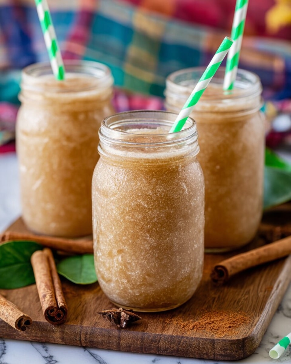 The image shows three clear glass jars filled with a smooth, light brown slushy drink that has a slightly frosty texture. Each jar has a green and white striped paper straw sticking out from the top. The jars are placed on a wooden board with cinnamon sticks and green leaves scattered around. The background includes a colorful plaid cloth and a white marbled surface underneath. Photo taken with an iphone --ar 4:5 --v 7