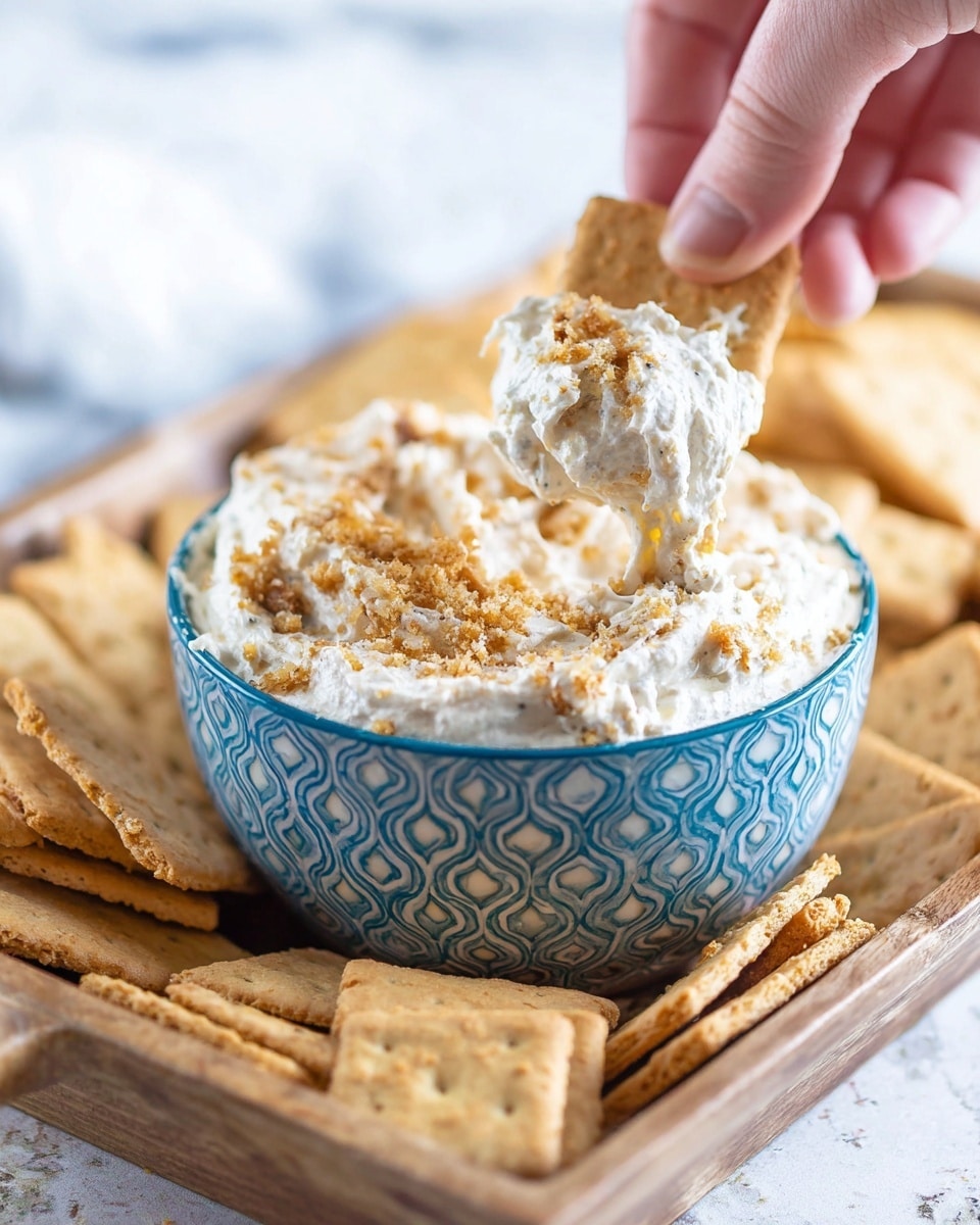 There is a small bowl with a blue and white geometric pattern filled with a creamy white dip that has small brown crumbs mixed in and sprinkled on top. The bowl is placed in a wooden tray filled with many rectangular, light brown crackers. A woman's hand is dipping one cracker into the mixture, showing the thick and textured cream clinging to it. The background has a white marbled texture. photo taken with an iphone --ar 4:5 --v 7