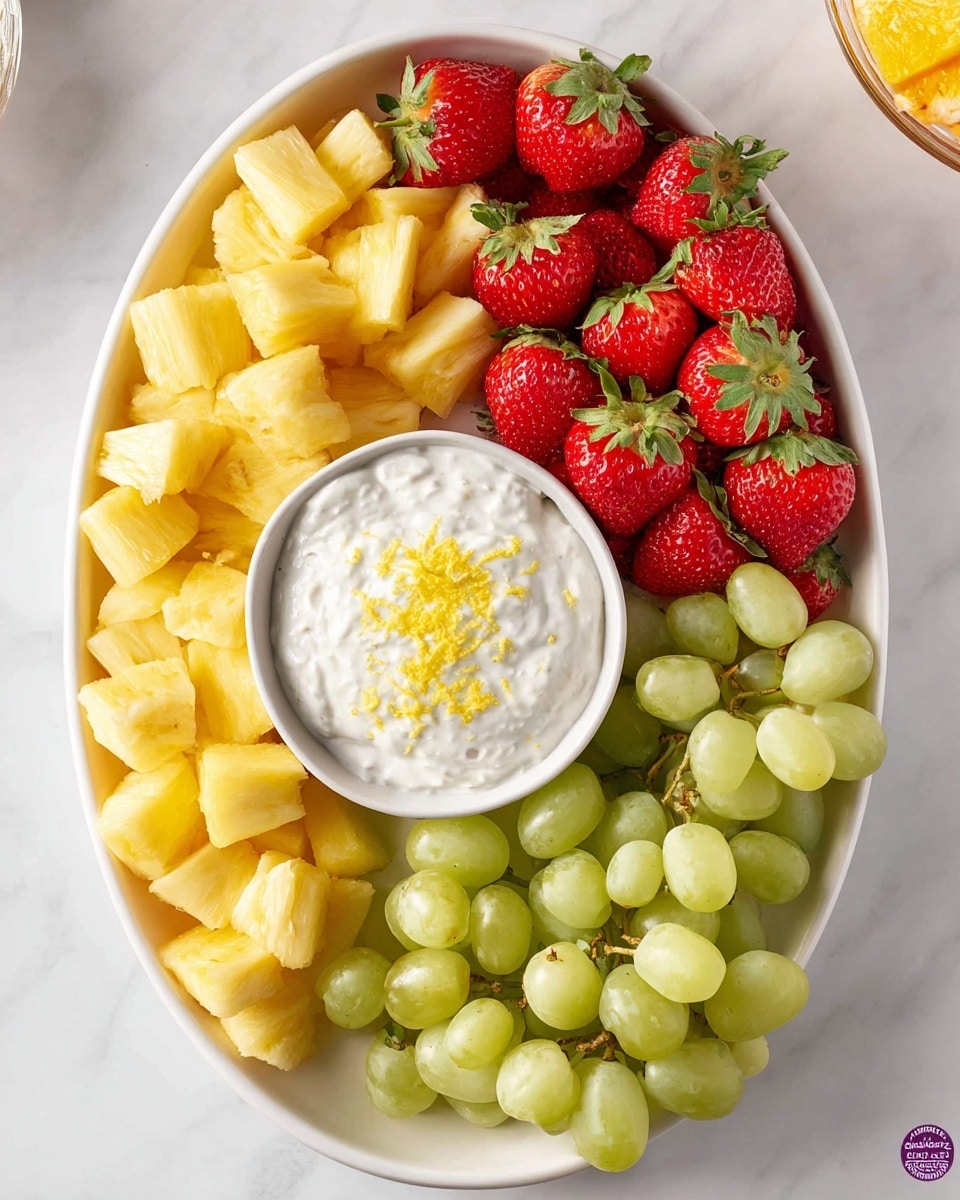 An oval white plate holds three groups of fresh fruit arranged around a small round white bowl of creamy white dip sprinkled with bright yellow zest. On the left side are chunks of yellow pineapple with a juicy texture, at the top are vibrant red strawberries with green leafy tops, and on the right side are plump light green grapes with smooth skins. The plate sits on a white marbled surface. photo taken with an iphone --ar 4:5 --v 7