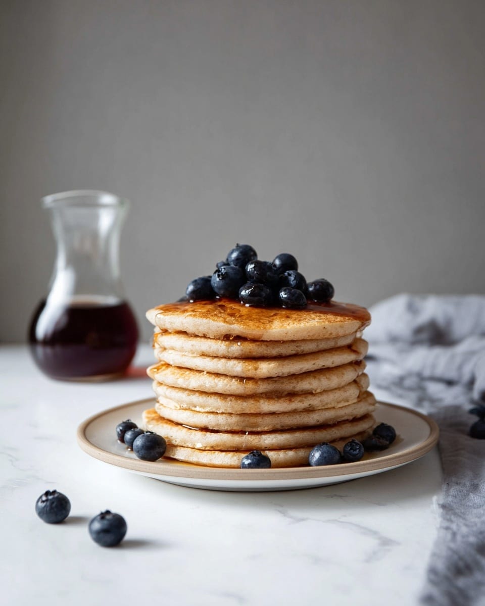 A stack of seven light brown pancakes sits on a white plate with a slightly raised rim, placed on a white marbled surface. The pancakes are evenly layered, with syrup drizzled lightly over the top and a cluster of fresh blue-black blueberries crowning the stack. Several blueberries are scattered on the plate around the base of the pancakes. In the background to the left, there is a clear glass pitcher holding dark syrup. The plain gray wall behind creates a simple, clean backdrop. Photo taken with an iphone --ar 4:5 --v 7