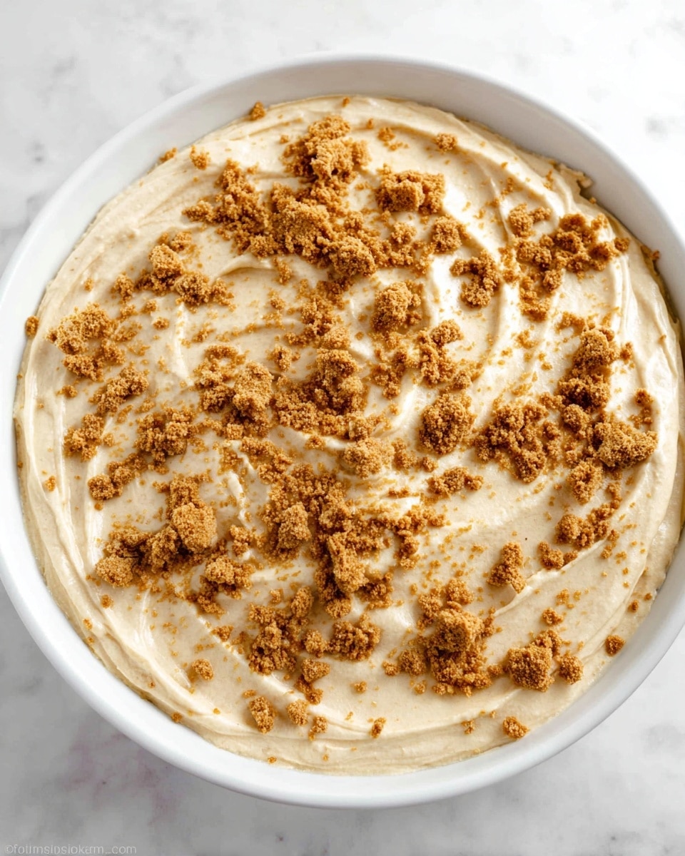 A white dish holds a creamy beige layer of smooth, thick dessert mixture spread evenly across the top. On this top layer, light brown crunchy crumbs are scattered unevenly, adding texture contrast. The creamy layer has soft swirls and slight peaks, showing it was spread with a utensil. The dish is set on a white marbled surface. photo taken with an iphone --ar 4:5 --v 7