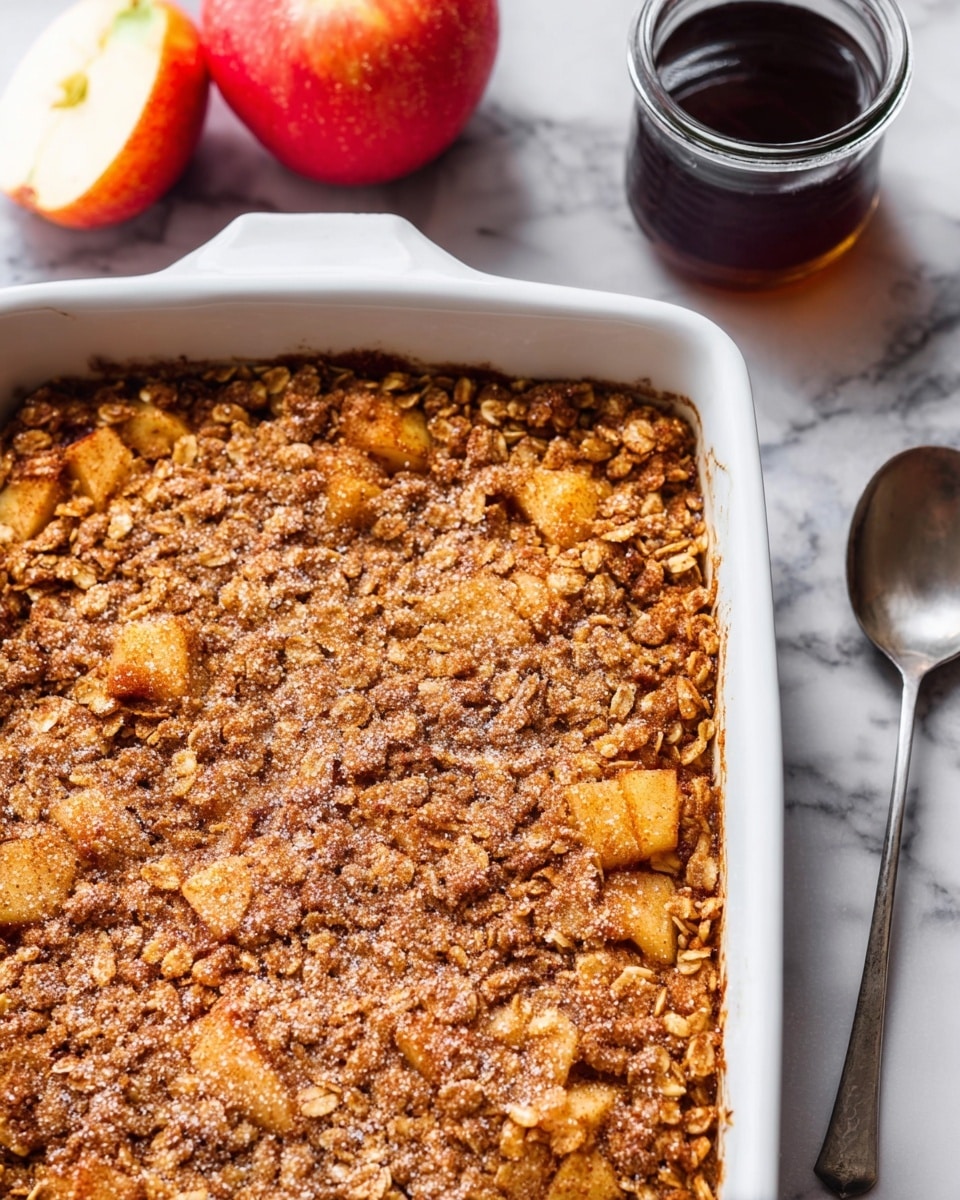 A rough-textured, golden brown baked oat square on a white plate with visible oats and small chunks of light apple pieces, sprinkled lightly with powdered sugar. The square sits next to a vintage silver fork placed diagonally across the plate, with a small drizzle of honey near the bottom right. A glass of amber-colored liquid and a red apple with a white interior slice are blurred out in the white marbled textured background. photo taken with an iphone --ar 4:5 --v 7
