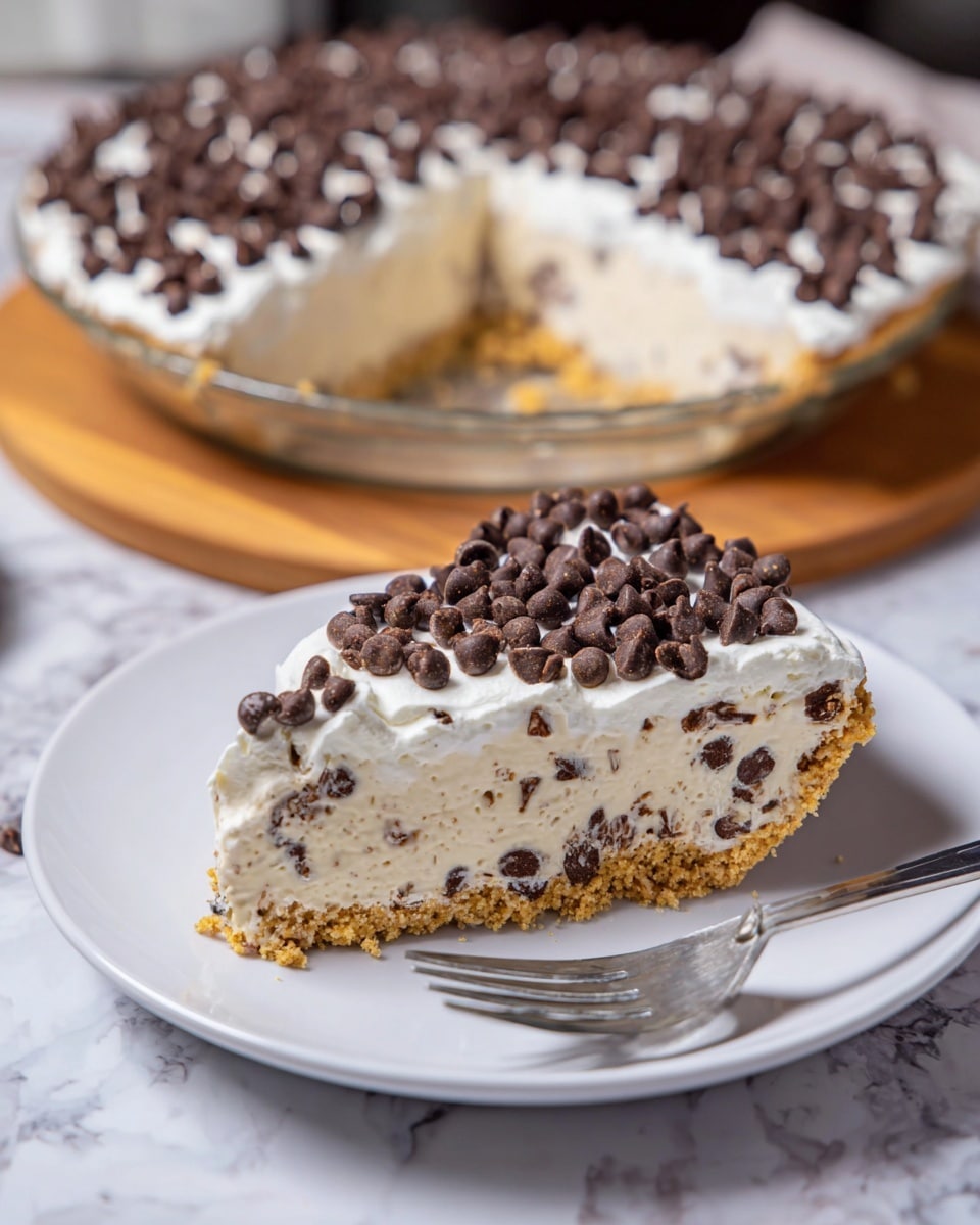 A slice of pie is shown on a white plate resting on a wooden board with a fork beside it. The pie slice has three main layers: the bottom layer is a golden brown crumbly crust, the thick middle layer is a creamy, light beige filling with small chocolate chips mixed inside, and the top layer is a generous spread of white cream dotted with more chocolate chips. The whole pie in the background is on a white plate, matching the small plate, and the top edge is covered with a ring of chocolate chips. The surface beneath everything is a white marbled texture. photo taken with an iphone --ar 4:5 --v 7
