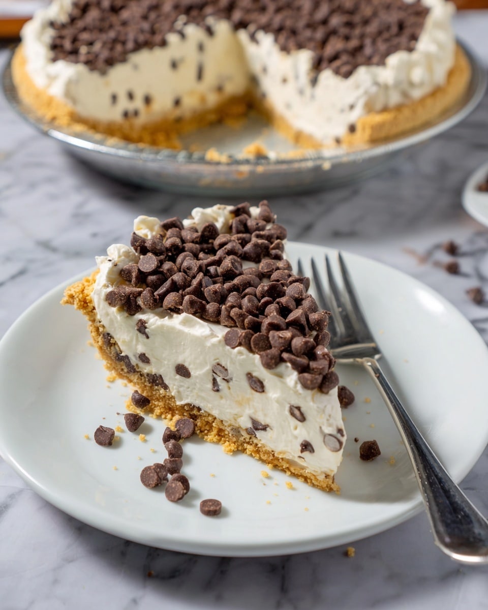 The image shows a slice of creamy dessert pie on a white plate with a silver fork beside it. The pie has three clear layers: a crumbly golden crust at the bottom, a thick creamy white middle layer dotted with small chocolate chips, and a topping of whipped cream covered with a dense layer of chocolate chips. Some chocolate chips and crumbs are scattered around the slice on the plate. In the background, the rest of the pie is visible on a white plate, showing the same layered texture and chocolate chip topping. The whole setup is placed on a white marbled surface. photo taken with an iphone --ar 4:5 --v 7