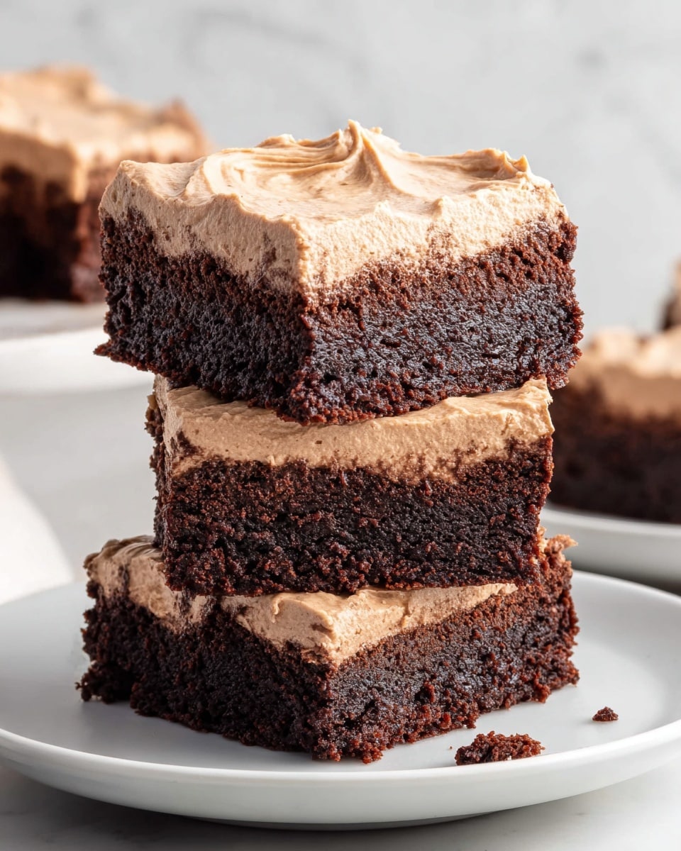 Three square pieces of chocolate brownies are stacked on a white plate, each with two layers: a dense, dark brown base with a rich, moist texture, and a thick, light brown, creamy frosting on top that looks soft and airy with gentle swirls. The edges of the brownies are slightly crumbly, and there are a few small crumbs scattered on the plate. The background shows a white marbled texture with soft natural light coming from the side. Photo taken with an iphone --ar 4:5 --v 7