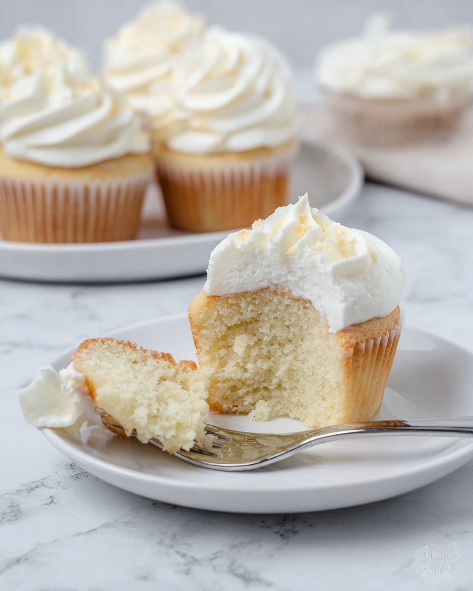 The image shows several vanilla cupcakes arranged on a white plate with a white marbled background. Each cupcake has one main layer of light golden cake topped with a thick swirl of smooth, creamy white frosting that forms a soft, wavy spiral pattern. The frosting is shiny and looks soft with a glossy texture. The cupcakes are close together and fill most of the plate, creating a neat, uniform look. Photo taken with an iphone --ar 4:5 --v 7