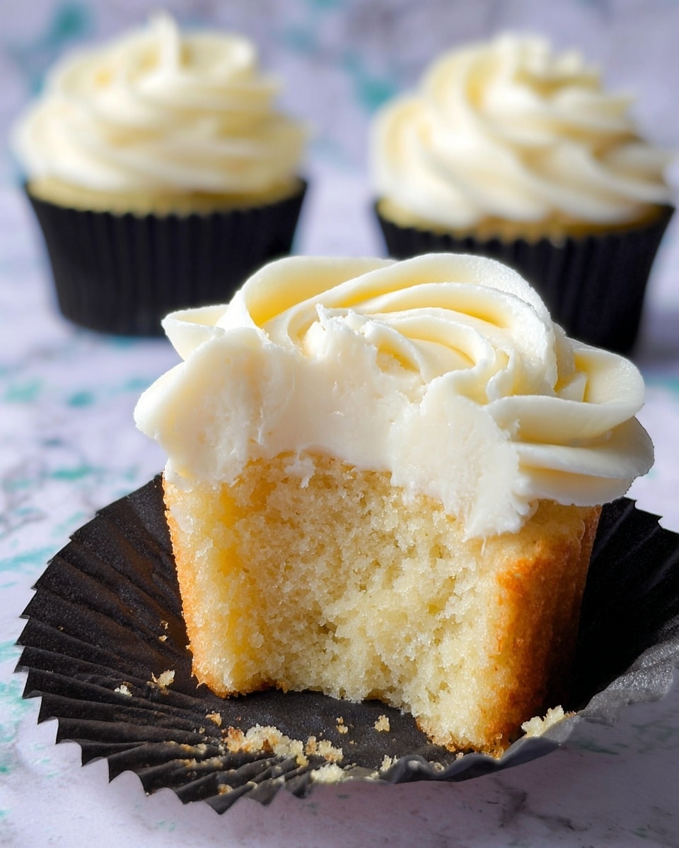 A close-up shot of a cupcake with two layers visible: the bottom layer is a soft, light yellow sponge cake with a moist and crumbly texture, while the top layer is thick, creamy white frosting swirled in a rose-like pattern. The cupcake wrapper is black and slightly open, showing the bitten edge of the cake. In the blurred background, two more cupcakes with the same frosting and black wrappers are displayed on a surface with a white marbled texture. photo taken with an iphone --ar 4:5 --v 7