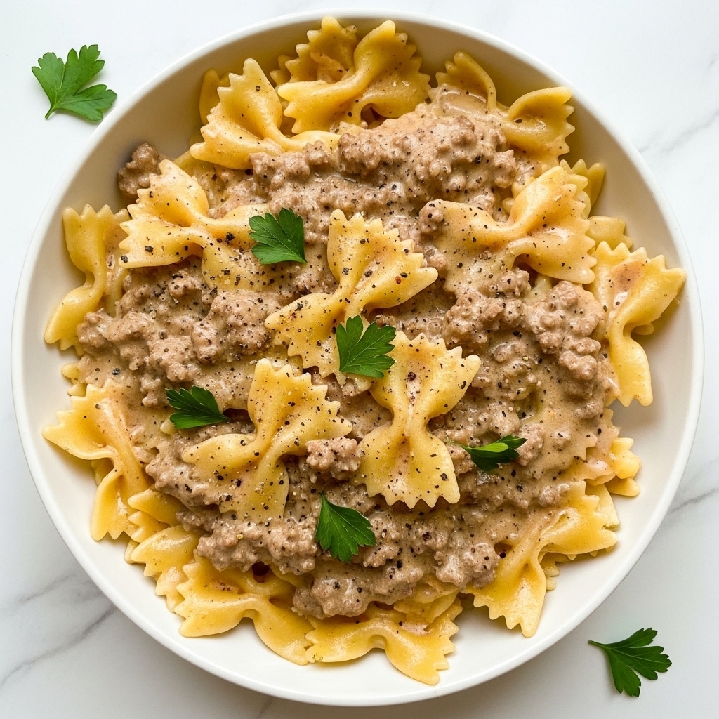 A white bowl filled with farfalle pasta covered in a creamy, light brown meat sauce with small bits of ground beef spread evenly throughout. The farfalle pasta pieces are golden-yellow with ruffled edges, slightly glossy from the sauce that coats them. The meat sauce looks rich and smooth, with visible specks of black pepper scattered on top. Small green parsley leaves are placed on top and around the dish for garnish. The bowl sits on a white marbled surface, highlighting the warm colors of the food. photo taken with an iphone --ar 4:5 --v 7
