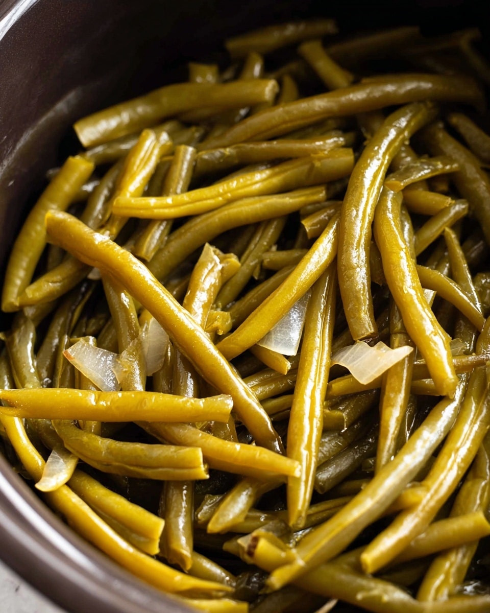 The image shows a close-up of cooked green beans inside a dark pot. The beans are long and slim with a shiny, slightly oily surface indicating they have been cooked well. There are small pieces of translucent, soft onions scattered among the green beans. The green beans are mostly a muted yellow-green color, with some parts showing a subtle brown tint from cooking. The texture looks tender and moist, with the beans overlapping in a loose pile within the pot. The background around the pot is a white marbled texture. photo taken with an iphone --ar 4:5 --v 7