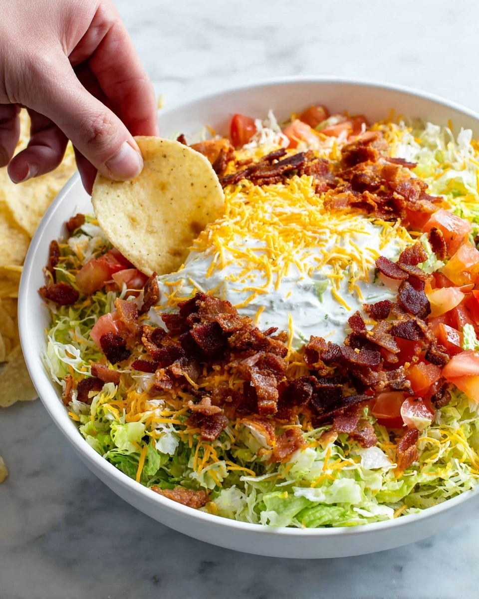 A white bowl filled with a dip that has four main visible layers: a bottom layer of green shredded lettuce around the edges, a middle layer of orange and red diced tomatoes and crispy dark brown bacon pieces scattered on top, then a middle-to-top layer of white creamy sour cream in the center, partially covered with more shredded light yellow and white cheese, and a pale yellow round chip dipped into the sour cream layer held by a woman's hand from the right side. The bowl sits on a white marbled surface. Photo taken with an iphone --ar 4:5 --v 7
