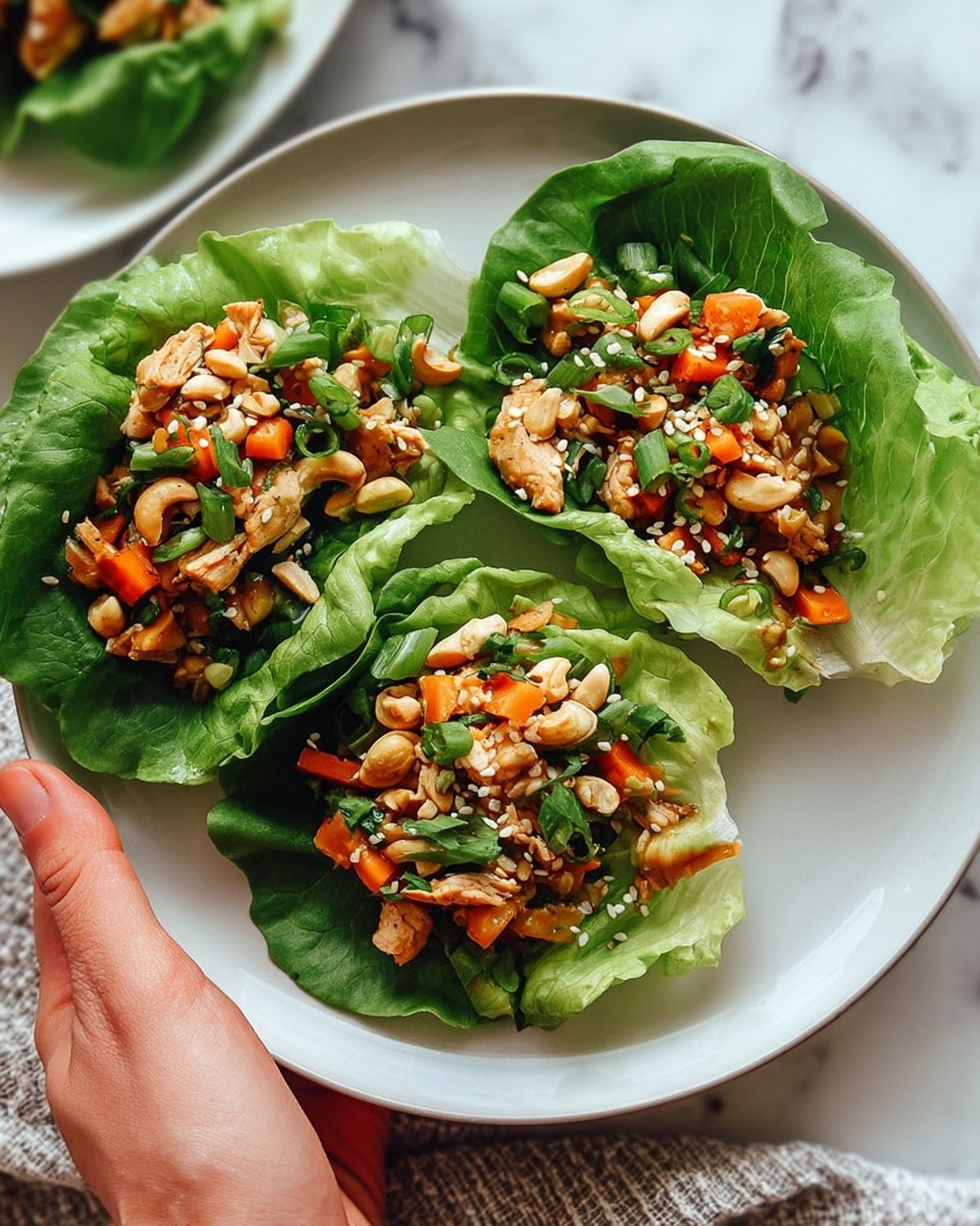 The image shows three fresh green lettuce cups placed on a white plate with a slight rim. Each lettuce cup acts as a bowl, holding a colorful mix of ingredients. The bottom layer is crisp green lettuce, layered with a vibrant mixture of cooked chicken pieces, small orange carrot cubes, green chopped herbs, and green onions. This filling is topped with light brown cashew nuts and sprinkled with white sesame seeds. A woman's hand is holding the edge of the plate on the right side. The plate is set on a white marbled surface with a hint of a textured fabric at the bottom left corner. photo taken with an iphone --ar 4:5 --v 7