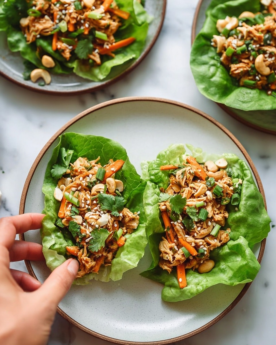 Three lettuce wraps are placed on a white plate with a light brown rim, set on a white marbled surface. Each wrap has one large bright green leafy lettuce base, topped with shredded light brown chicken mixed with small orange carrot sticks, chopped green onions, and sprinkled with chopped cashews and white sesame seeds. A few cilantro leaves add extra green color for garnish. A woman's hand is gently touching one of the wraps from the side. In the background, two more plates with similar wraps are partially visible. Photo taken with an iphone --ar 4:5 --v 7