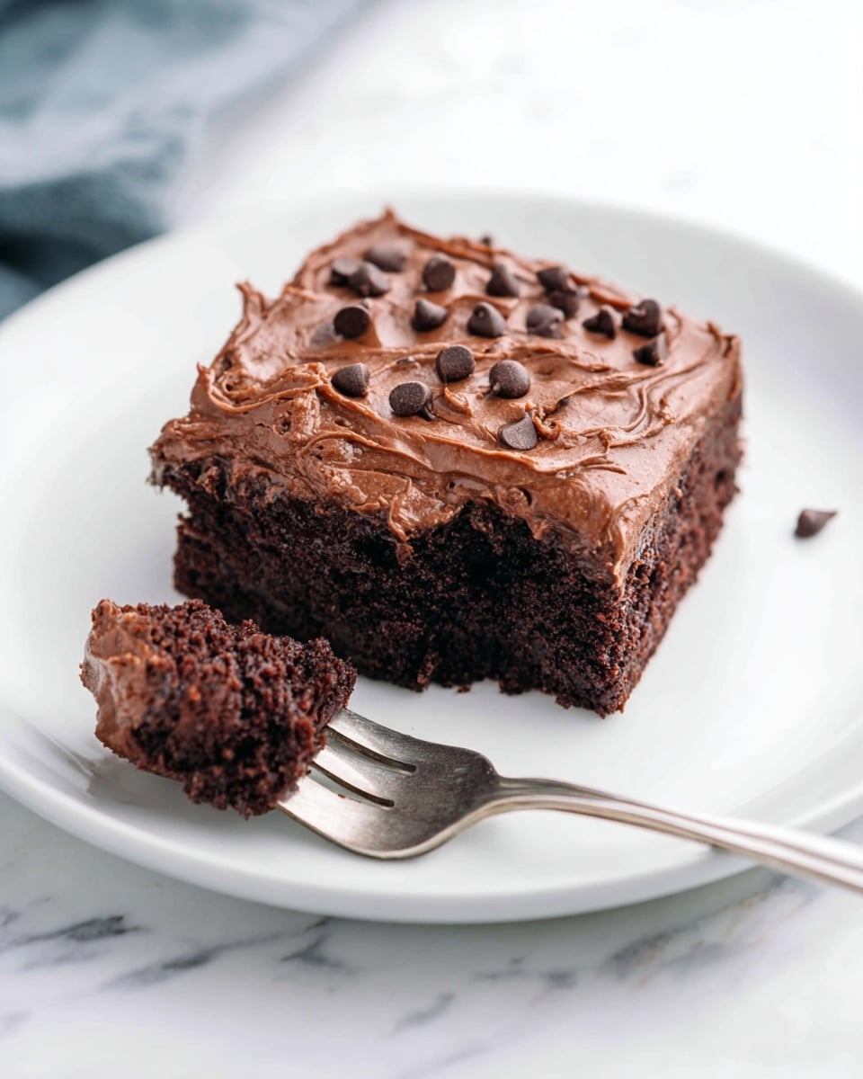 A square piece of chocolate cake sits in the center of a white plate on a white marbled surface; the cake has two layers, a dark moist chocolate base and a thick, smooth layer of chocolate frosting on top, decorated with scattered small chocolate chips; a silver fork holds a bite-sized piece of the cake with visible creamy frosting and crumbly cake texture. photo taken with an iphone --ar 4:5 --v 7