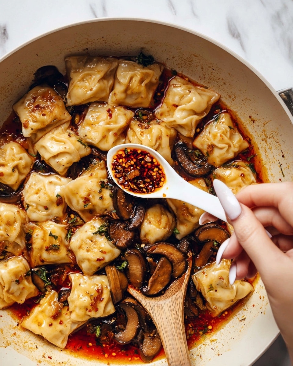 A white pan filled with small, square dumplings that have soft, pale yellow skins, cooked with dark brown, sliced mushrooms. These sit in a rich, oily red sauce that has flecks of chili flakes visible throughout. A wooden spoon rests on the right edge of the pan, and a woman's hand with long nails is holding a white spoonful of chunky chili oil above the dumplings. The background is a white marbled texture. photo taken with an iphone --ar 4:5 --v 7