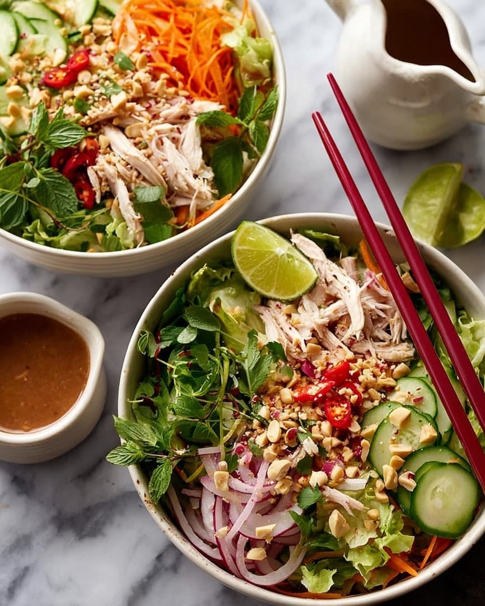 A large silver bowl filled with a colorful layered salad sitting on a white marbled surface; the bottom layer is a mix of shredded green lettuce and sliced cucumbers creating a fresh green base, topped with thin strips of red bell pepper and shredded pale chicken pieces scattered evenly, followed by slivers of red onion and bright orange carrot shreds adding contrast; a sprinkle of crushed peanuts and chopped fresh green herbs like cilantro and mint is piled higher in the center, with three lime wedges placed on the right side on top of the salad for a vibrant touch; a wooden spoon rests inside the bowl; behind the bowl is a small white gravy boat filled with brown dressing and some green herbs scattered loosely around; photo taken with an iphone --ar 4:5 --v 7