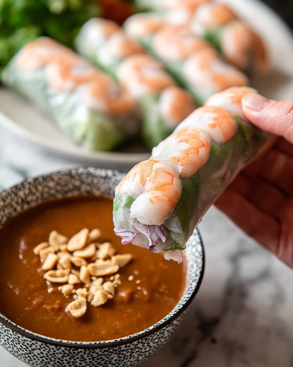A close-up of a woman's hand holding a translucent shrimp spring roll. The roll has three visible layers: a thin, clear rice paper outer layer showing orange and white shrimp pieces arranged on the outside, fresh green leafy herbs under the shrimp, and a light purple or white vegetable layer at the base inside. In the background, several more spring rolls rest on a white plate placed on a white marbled surface. In front, a patterned white bowl filled with thick, reddish-brown peanut sauce topped with chopped peanuts is partially visible. Photo taken with an iphone --ar 4:5 --v 7