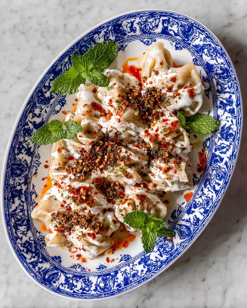 The dish is a white oval plate with a classic blue patterned rim, filled with about ten dumplings or rolls made from light beige dough, each folded into a pouch shape. These dumplings are layered with a thick white yogurt or cream sauce that covers most of their surface, sprinkled generously with dark green dried herbs and reddish-orange chili flakes and oil drizzled unevenly. There are two fresh mint leaves placed on the side of the plate as garnish. The plate sits on a white marbled surface. photo taken with an iphone --ar 4:5 --v 7