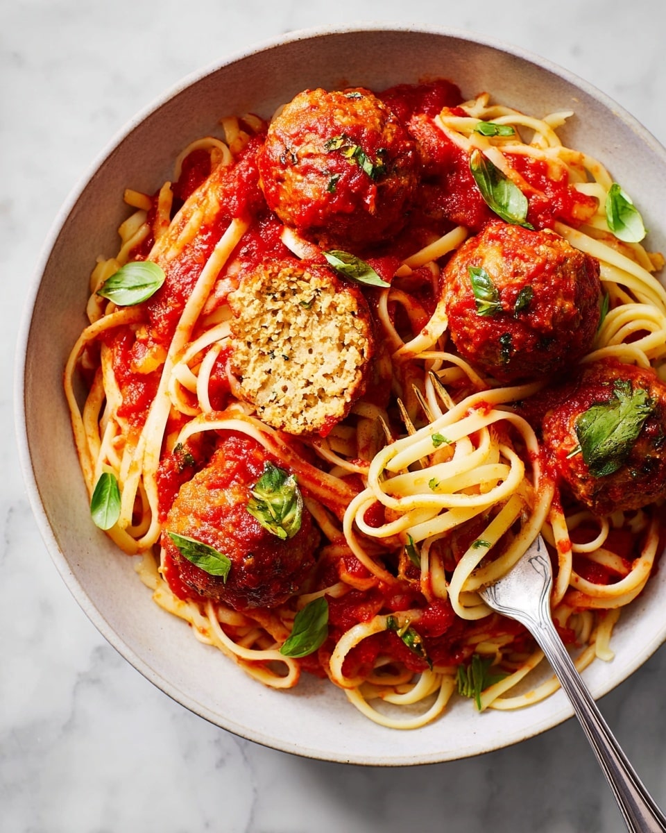 A close-up of a white shallow bowl filled with three layers: first, thick red tomato sauce covers the bottom; second, light yellow pasta noodles are twisted and mixed throughout; third, round, brownish-red meatballs coated in sauce are placed on top, one of which is cut open showing a crumbly, textured inside with bits of herbs. Scattered green basil leaves add contrast. A silver fork holds a small bundle of noodles near the bottom right of the bowl. The dish is on a white marbled surface. photo taken with an iphone --ar 4:5 --v 7