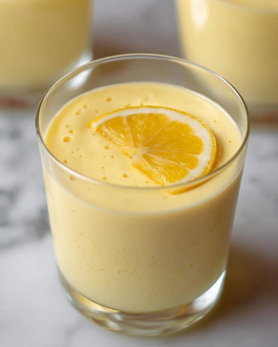 The image shows a close-up of a clear glass filled with a smooth, creamy yellow dessert, likely a lemon mousse or custard. The dessert fills almost the entire glass, with small bubbles visible on its surface, adding texture. On top, there is a thin, bright yellow lemon slice with a slightly glossy look, resting flat and centered as the only garnish. The glass is placed on a white marbled background, highlighting the soft yellow color of the dessert. Another similar glass is partly visible blurred in the background. Photo taken with an iphone --ar 4:5 --v 7