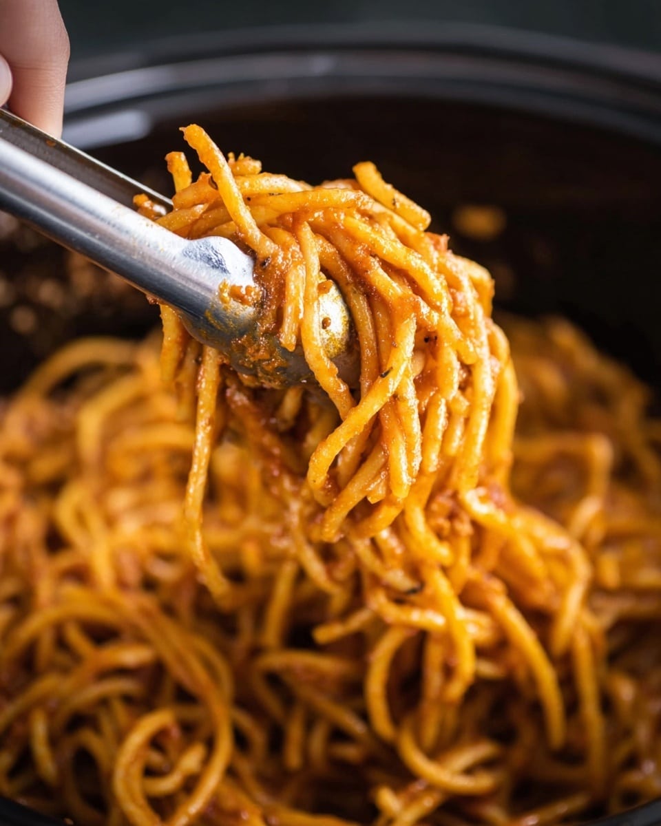 A close-up view of cooked spaghetti noodles coated in a reddish-orange sauce with visible small bits of seasoning and texture on each strand, lifted up by shiny silver metal tongs held by a woman's hand on the left side; the noodles are thick and intertwined with a slightly oily and seasoned surface, all seen against a blurred dark pot background. photo taken with an iphone --ar 4:5 --v 7