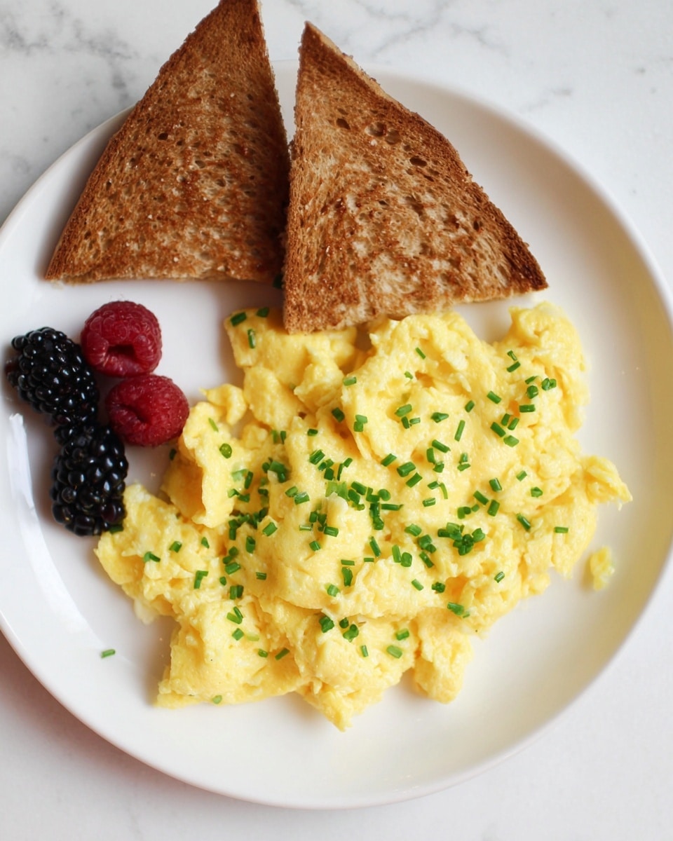 The image shows a white plate with three main parts: a large scoop of soft, fluffy yellow scrambled eggs sprinkled with small pieces of chopped green chives on the right side, two triangular slices of browned toast with a slightly crunchy texture placed at the top left, and a small cluster of mixed berries including blackberries and raspberries at the bottom left. The plate sits on a white marbled surface that adds a clean and bright background to the dish. Photo taken with an iphone --ar 4:5 --v 7