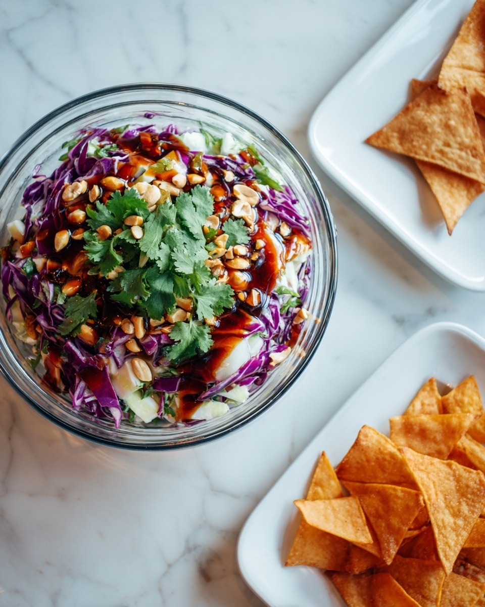 A clear glass bowl is placed on a white marbled surface, filled with a colorful layered salad. The bottom layer looks light and creamy, topped with chopped fresh herbs scattered evenly. Over this is a layer of thin purple cabbage strips mixed with small green herb leaves and crushed peanuts spread across the top. A dark brown sauce is drizzled in thin lines over the entire salad. Next to the bowl, on the left, is a white plate holding several triangular crispy golden-brown chips stacked on each other. The scene is bright and fresh. Photo taken with an iphone --ar 4:5 --v 7